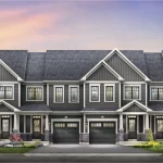 Row of newly designed townhouses at Empire Legacy with dark grey vinyl siding, integrated front-garage doors, and small landscaped front yards under a twilight sky.