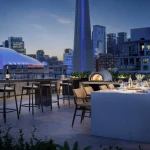 Outdoor amenity terrace at Q Tower featuring a long dining table, pizza oven, and barbecue station with scenic evening views of the CN Tower and illuminated Rogers Centre.