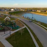 Aerial view of the Wildflower master-planned community showing a modern playground park, walking paths, a scenic stormwater pond, and surrounding residential neighborhoods under a clear sky.