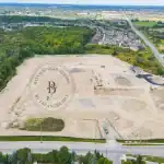 Aerial drone view of the Bayview Trail construction site in Aurora, showing land grading, heavy machinery, and proximity to a lush green forest and existing residential neighborhoods.
