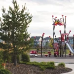 A modern playground in Cy Becker park featuring a unique red and black climbing structure with a silver slide, surrounded by green grass, trees, and residential houses in the background.