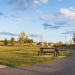 A paved walking path winding through a green park in the Cy Becker community, lined with picnic tables, young trees, a lamppost, and rolling grassy hills under a blue sky.
