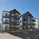 Exterior view of modern multi-story condominium buildings at Desrochers Villages, featuring a mix of dark grey vertical siding, white panels, warm wood accents, and private balconies under a clear blue sky.