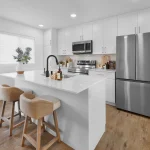 Bright, open-concept kitchen interior at Desrochers Villages with a white quartz island, black gooseneck faucet, tan counter stools, white cabinetry, and stainless steel appliances.