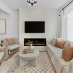 Minimalist modern living room interior at Desrochers Villages featuring a wall-mounted flat-screen TV above a linear fireplace, an off-white sofa with tan throw pillows, and two rattan accent chairs.