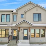 Exterior of modern townhomes at Desrochers Villages in the winter, showing light beige vertical siding, dark window frames, and snow-covered steps leading to front doors.