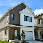 Exterior of the Aspen detached home model in the Aster community, featuring a modern two-story design with white and brown vertical siding, a single-car garage, and a bright blue sky background.