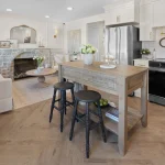 Open-concept main floor interior of the Mila townhouse at Marquis Meadows, featuring a modern kitchen with stainless steel appliances, a wooden island with barstools, herringbone flooring, and a cozy stone fireplace in the living area.