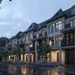 Evening streetscape of Fifth Avenue Homes in Woodbridge, showing modern luxury townhouses with illuminated large windows, stone pillars, and professional landscaping on a rainy night.