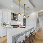 Modern luxury kitchen at Fifth Avenue Homes Woodbridge with white cabinetry, a large waterfall island, gold hardware, and high-end stainless steel appliances.