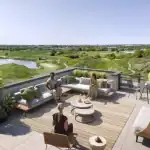 Luxury rooftop patio at Spruce Trails featuring modern outdoor furniture, a sectional sofa, and a scenic panoramic view of rolling green hills, a pond, and the city skyline in the distance.