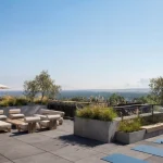 Rooftop patio amenity space at Black Birch Heights Condos featuring outdoor lounge seating, dining areas under umbrellas, concrete planters with greenery, and yoga mats looking out over a scenic view.