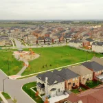 High-angle aerial view of a master-planned residential neighborhood in Mayfield Village, showcasing rows of modern suburban houses, paved streets, and a large central green park with a children's playground and gazebo under an overcast sky.