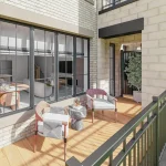 Private balcony rendering at OG Urban Towns featuring wood plank flooring, modern outdoor chairs, a small marble side table, and a large multi-pane glass window looking into the open-concept living area.