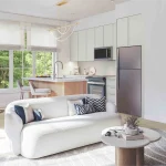 Bright, open-concept interior rendering of an OG Urban Towns unit, showcasing a modern kitchen with light cabinets and stainless steel appliances, a cozy dining area, and a contemporary living room with a white sofa.