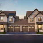 Front exterior view of a semi-detached home block at Harwood Place, featuring dual-car garages, mixed stone and vertical siding facades, covered entryways, and warm interior lighting at twilight.