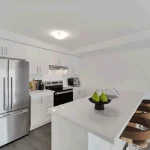 Modern kitchen interior at Countryside Urban Towns with white cabinetry, stainless steel appliances, a kitchen island, and barstool seating.