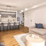 Contemporary open-plan living room and kitchen design at Sorella Towns featuring grey cabinetry, modern island bar stools, a beige sectional sofa, light wood flooring, and neutral decor.