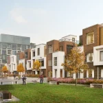 Street-level view of modern brick and white panel townhomes at Elements Milton with residents walking dogs along a landscaped green pedestrian pathway.