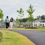 A couple holding hands walks along a paved pathway in a modern suburban neighborhood with newly built homes and a playground in Mount Pleasant West.