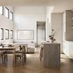 Modern open-concept kitchen and dining area at VALO showcasing minimalist white cabinets, a travertine waterfall island, wooden dining set, light herringbone hardwood floors, and double-height ceilings.
